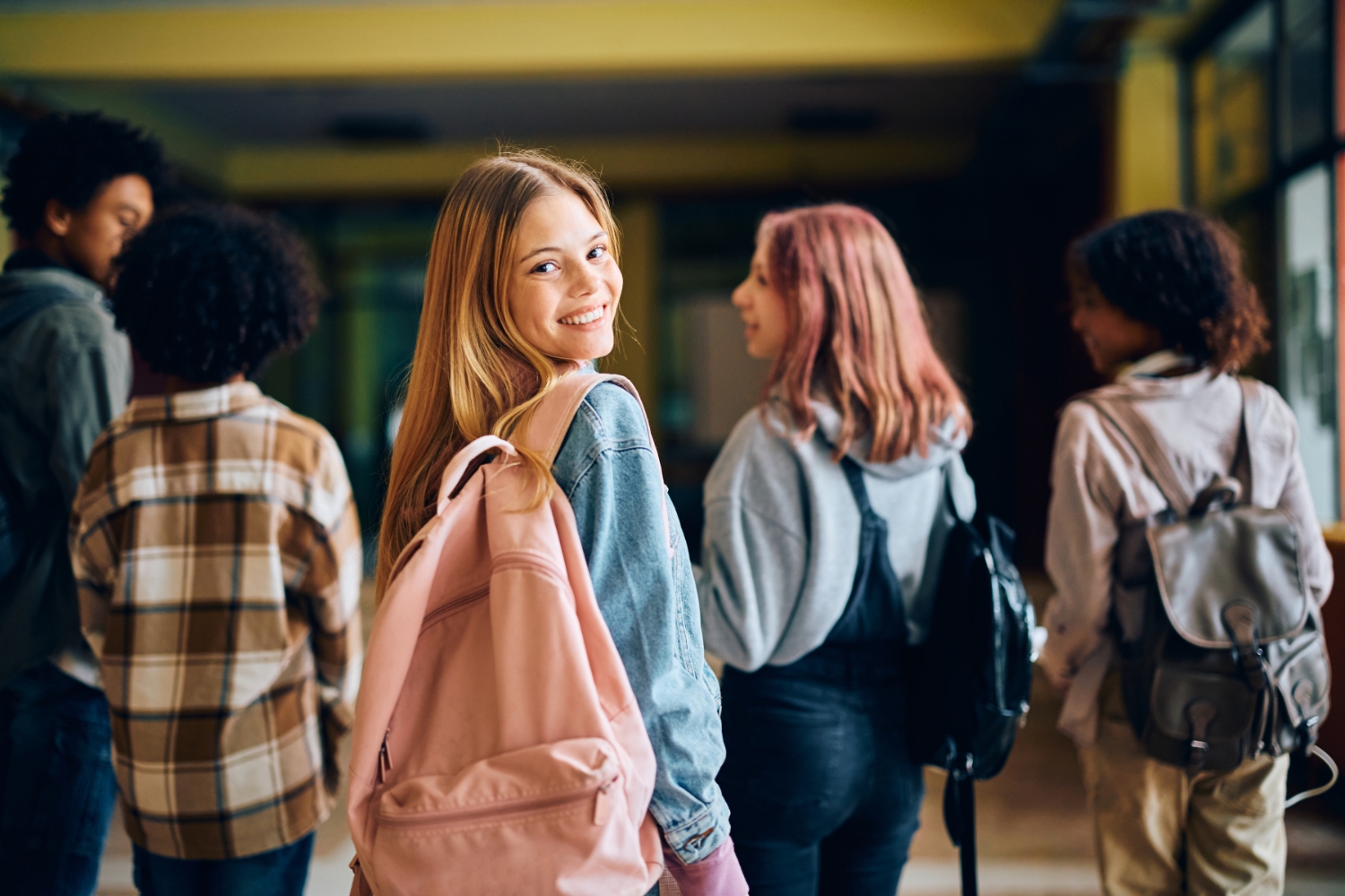 A group of teens walks down a school hallway with their backs turned, with one teen girl looking back and smiling at the camera