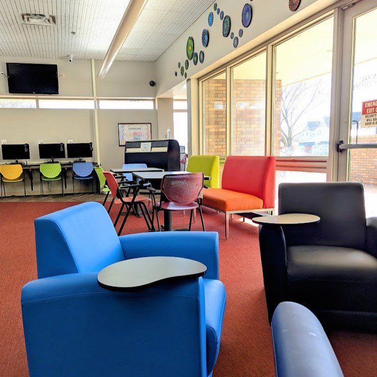 Two armchairms with fold-out desks. In the background are two tables with colorful benches on one side and chairs on the other.