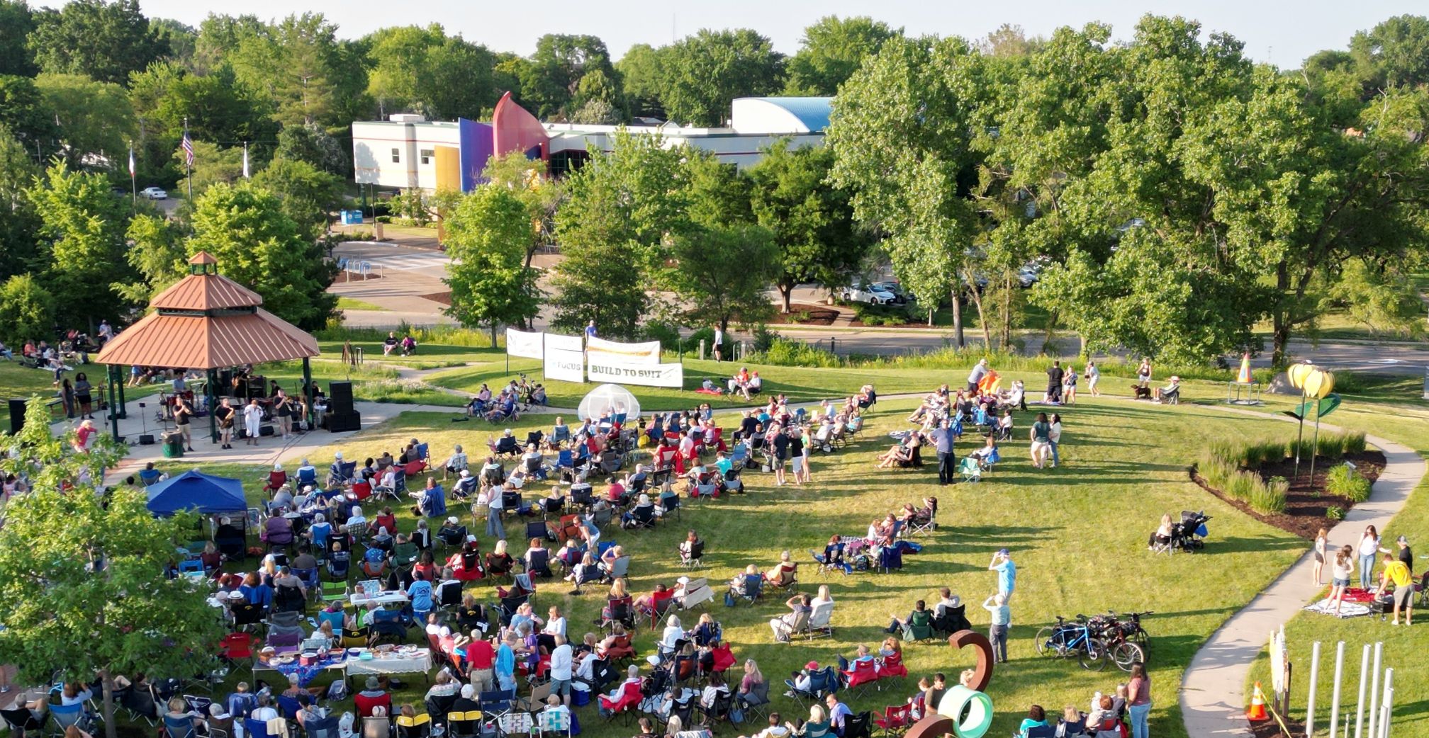 People in lawn charis in an open field around a gazebo, where a band performs. The Bettendorf Public Library is in the background.