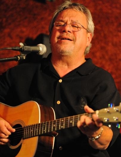 Middle-aged white male with gray hair and glassess sitting down and playing guitar