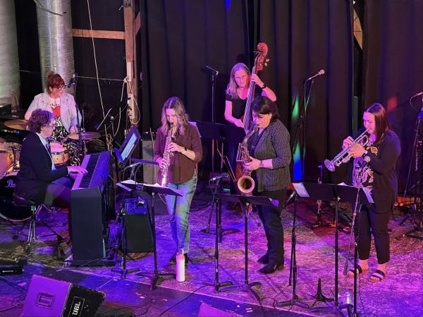 A group of six women playing various jazz instruments on a stage