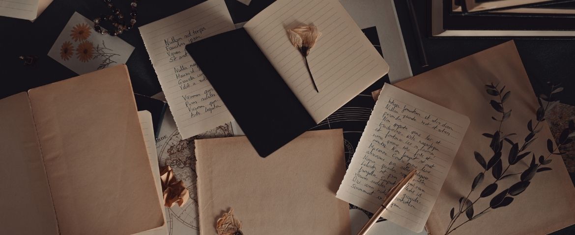 Blank paper and open notebooks with writing on them lay on a table scattered with dried flowers