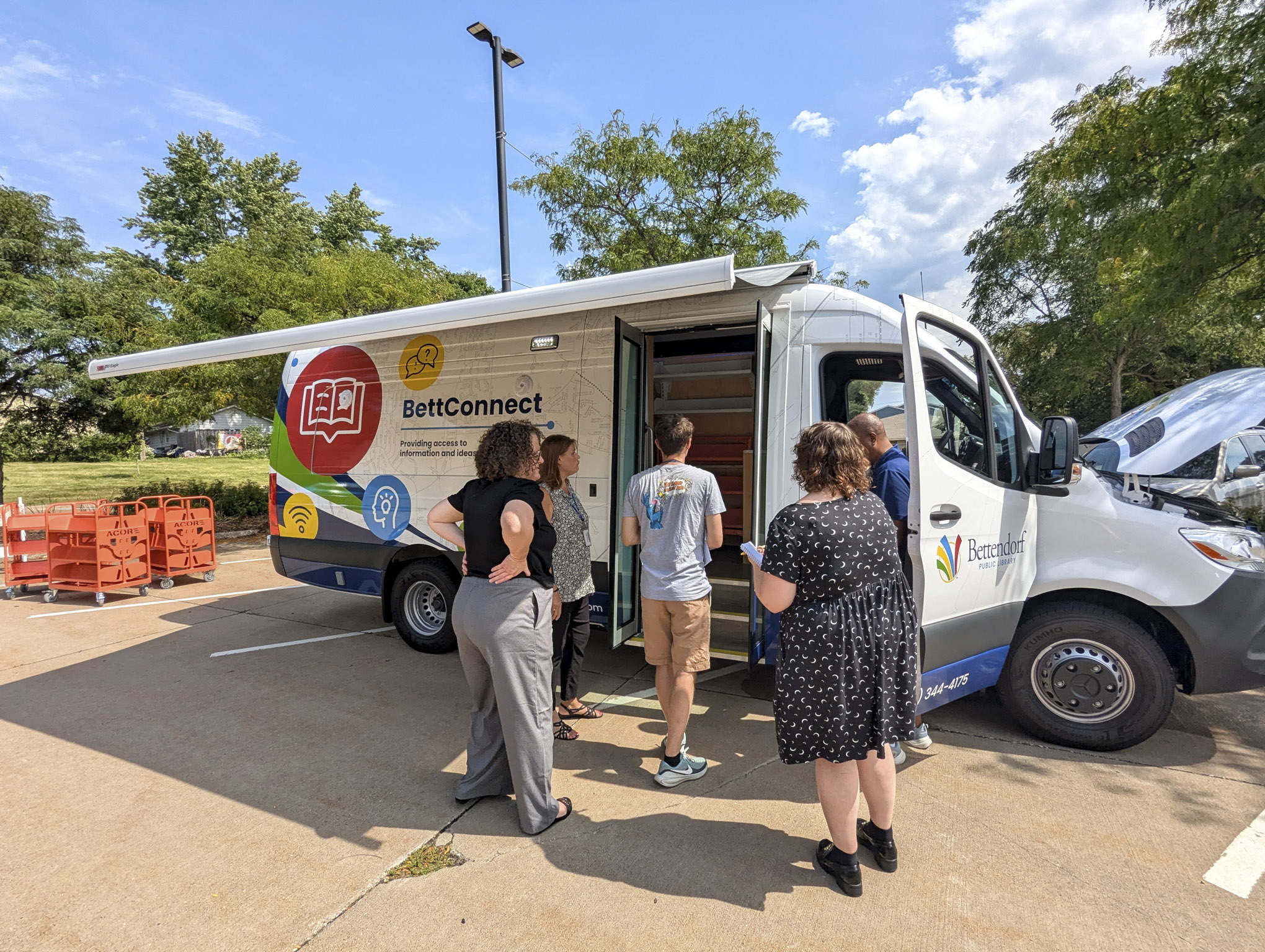 Library staff stands outside a library outreach vehicle van with an awning extended. The van is branded BettConnect and features colorful library icons.